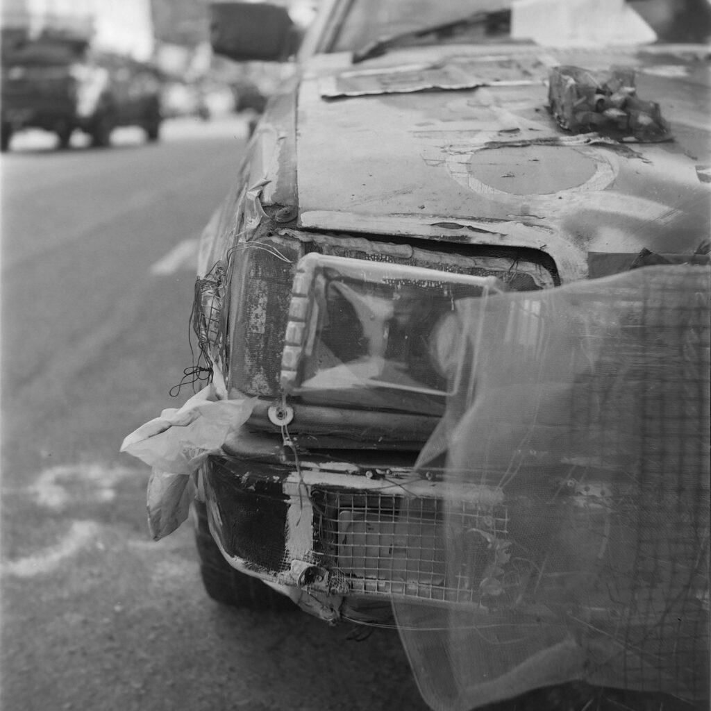 A close-up of a weathered car parked on an urban street, showing its damaged headlight and hood.